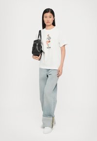 T-shirt blanc avec un imprimé graphique, jeans baggy bleu clair avec un accent jaune et un sac bandoulière texturé noir. Baskets blanches.