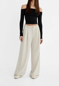 Zwarte off-shoulder top met lange mouwen, gecombineerd met lichtbeige wijde broek met voorplooien en een ceintuur in de taille.