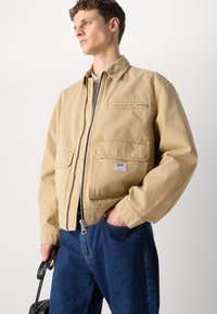 Beige bomullsjakke med glidelås foran, to store lommer og en glidelåslomme på brystet. Brukt over blå denim jeans.