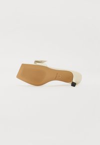 Beige high-heeled shoe with a glossy finish, pointed toe, bow accent on the strap, and a textured nude sole. Black heel detail.
