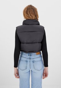 Woman with wavy light brown hair wearing a black puffer vest over a black long-sleeve top and light blue high-waisted jeans, facing away.