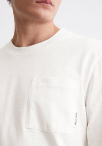 Weiße Baumwoll-T-Shirt mit einer Brusttasche. Der Stoff hat eine subtile Textur, mit einem Etikett am Rand der Tasche und einem runden Halsausschnitt.