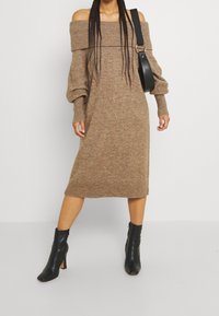 Strickkleid mit schulterfreier Form in Hellbraun, knielang mit langen Ärmeln. Verfügt über gerippte Bündchen und eine lockere Passform. Kombiniert mit schwarzen, hohen Ankle Boots.