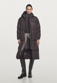 Long brown puffer coat with a zip front, hood, and quilted design. Paired with a gray sweater, belt, and black knee-high boots.