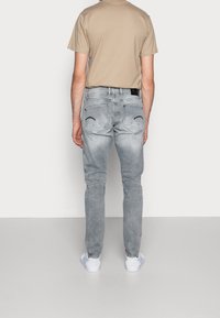 Jeans de mezclilla gris claro con bolsillos traseros que presentan un sutil pespunte curvado. Combinados con una camiseta beige y zapatillas blancas.