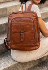 Ruskea nahkareppu, jossa on säädettävät hihnat, etutasku ja vetoketjusulkimet. Sileä pinta, jossa on ompeluyksityiskohtia ja ylhäältä oleva kahva.