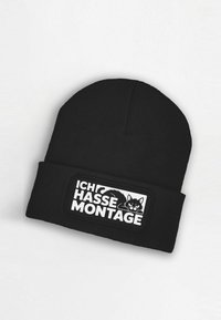 Black knitted beanie with a textured ribbed finish. Features a white patch with black text "ICH HASSE MONTAGE" and a cartoon cat illustration.