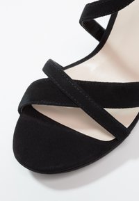 Sandale en suède noir avec des lanières croisées, une zone de orteil lisse et une semelle intérieure de couleur neutre. Le design met en avant une élégance minimaliste.