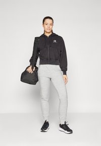 Svart cropped hettegenser med logo, grå joggebukser og svarte sneakers. En svart duffelbag holdes med den ene hånden. Jevne teksturer gjennom hele.