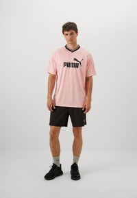 Šviesiai rožinė sportinė marškinė su juodais akcentais ir "PUMA" logotipu, derinama su juodomis šortais ir juodais sportiniai batais. Paprastas fonas.