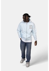 Felpa azzurra con zip e una grande lettera "C" nera sul petto, indossata sopra una t-shirt bianca e abbinata a jeans blu e sneakers grigie.