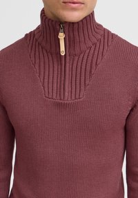 Gestrickter Pullover in dunklem Bordeauxrot, mit einem halben Reißverschlusskragen, der gerippte Textur aufweist, sowie einer Lederakzent am Reißverschluss.