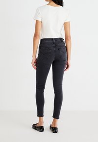 Jeans negros de corte alto y slim fit con un acabado mate. Incluyen dos bolsillos traseros y pasadores para cinturón, combinados con una camiseta blanca básica y zapatos negros planos.