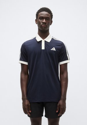 Navyblauw poloshirt met een crème-kleurige kraag en manchetten, met drie witte strepen op de schouders en een klein wit logo op de borst.