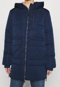 Granatowa ocieplana kurtka z zamkiem błyskawicznym, kapturem i czterema przednimi kieszeniami. Charakteryzuje się teksturowaną powierzchnią oraz luźnym krojem.