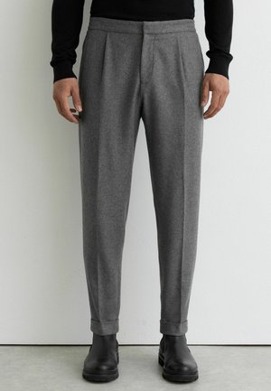 Homme portant un pantalon gris sur mesure avec des revers et des bottes en cuir noir, debout contre un mur blanc texturé.