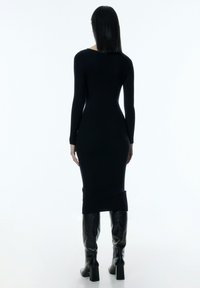 Schwarzes Strickkleid mit langen Ärmeln, figurbetontem Schnitt, geripptem Stoff und Schlitz im Rücken. Das Model trägt hohe schwarze Stiefel.