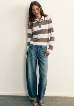 Frau steht vor einer schlichten Wand, trägt ein langärmeliges Poloshirt mit pinken und olivgrünen Streifen, weit geschnittene hochgekrempelte Jeans und pink karierte Schuhe.