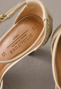 Beige vasket skindsandaler med en tan indersål, med et mærkelabel og justerbar ankelstrop. Tekstureret overfladedetalje synlig.