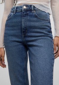 Högmidjade blå denimjeans med en klassisk passform, som har subtila slitningar och en knappstängning. Mjuk textur, fem synliga fickor.