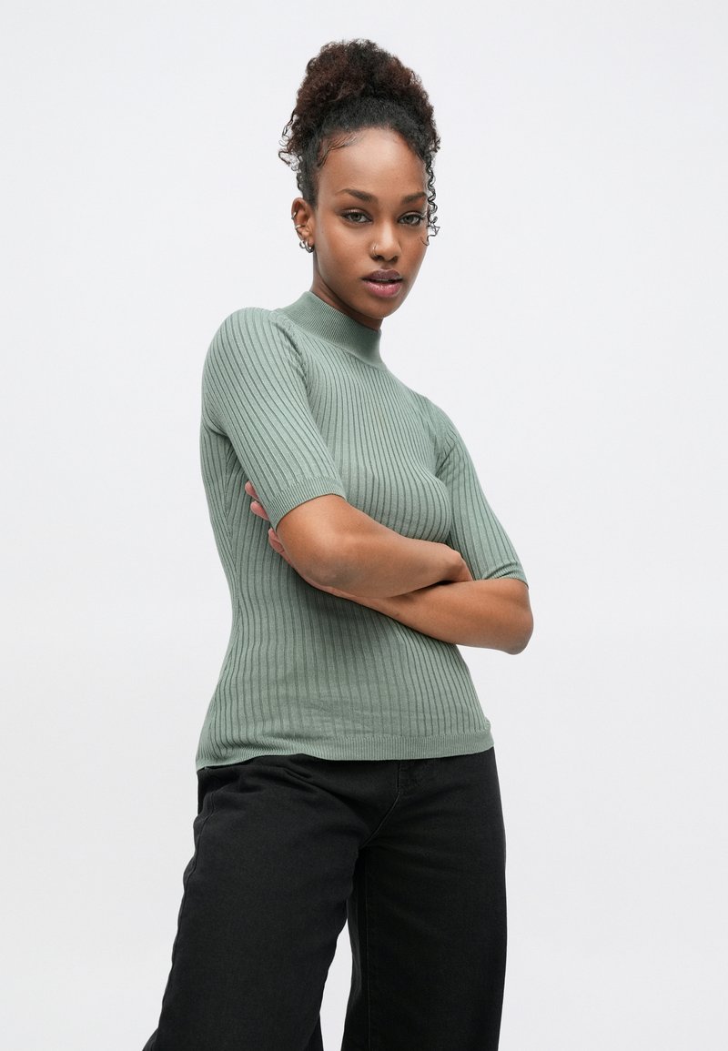 Πλεκτό πράσινο πουλόβερ με ριμπ ύφανση, ψηλό λαιμόκοψη και κοντά μανίκια. Έχει εφαρμοστή γραμμή και συνδυάζεται με μαύρο παντελόνι.