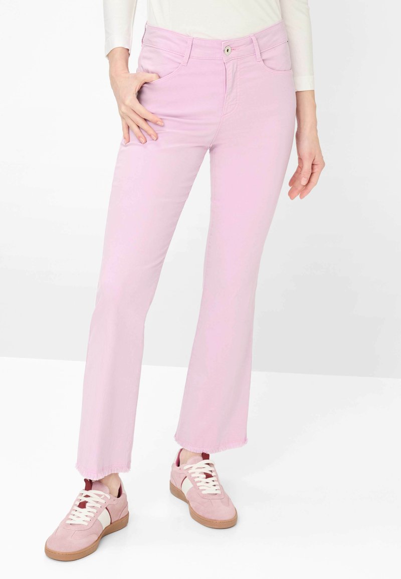 Vrouw draagt lichtroze cropped jeans met rafelige zomen en roze sneakers met witte veters en bruine zolen, staand met een hand in de zak.