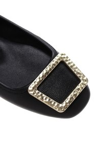 Chaussure plate en cuir noir avec un accent de boucle en or texturée. La chaussure présente un bout arrondi et une doublure intérieure lisse.