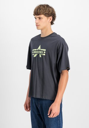 Tmav gray oversize tričko s krátkymi rukávmi, s svetlozeleným grafickým dizajnom so slovom "INDUSTRIES" v strede.