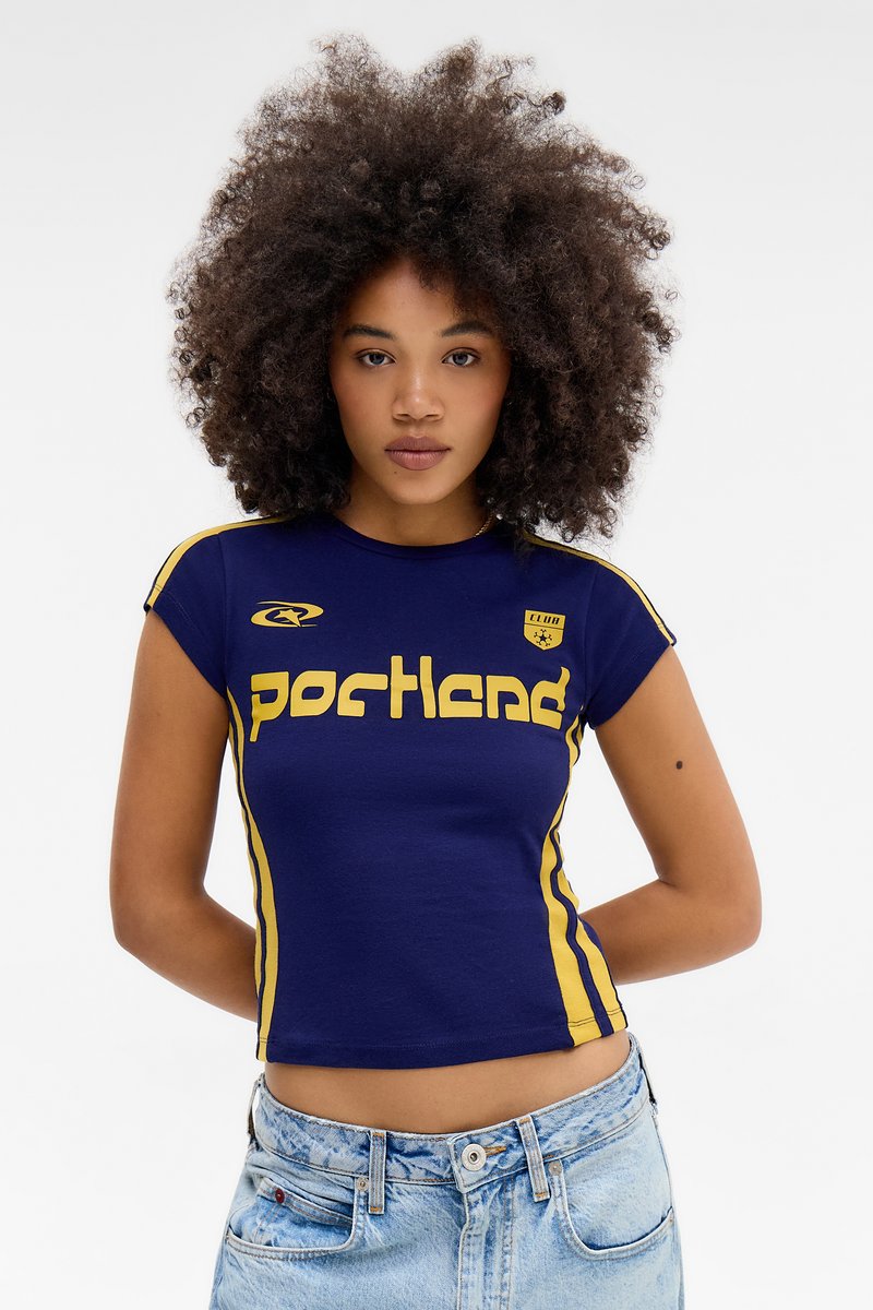 Tânără cu păr creț, purtând un tricou sportiv „Portland” albastru închis și galben și blugi din denim albastru deschis, stând în picioare pe un fundal simplu.