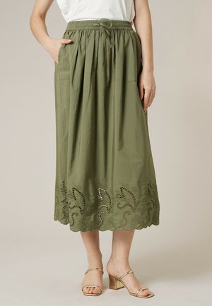 Femme portant une jupe midi vert olive avec un ourlet festonné brodé, une taille élastique, et des sandales à talons dorés, debout devant un fond uni.