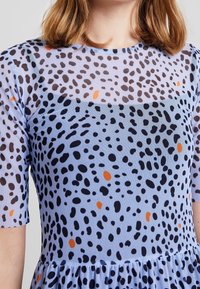 Průsvitné, světle modré šaty s černým a oranžovým skvrnitým vzorem, krátké rukávy a nařasená sukně na lemu.