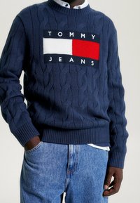 Mörkblå kabelstickad tröja med en kontrasterande rektangulär logotyp-patch med "TOMMY JEANS" i vita bokstäver och en block av röd och vit.