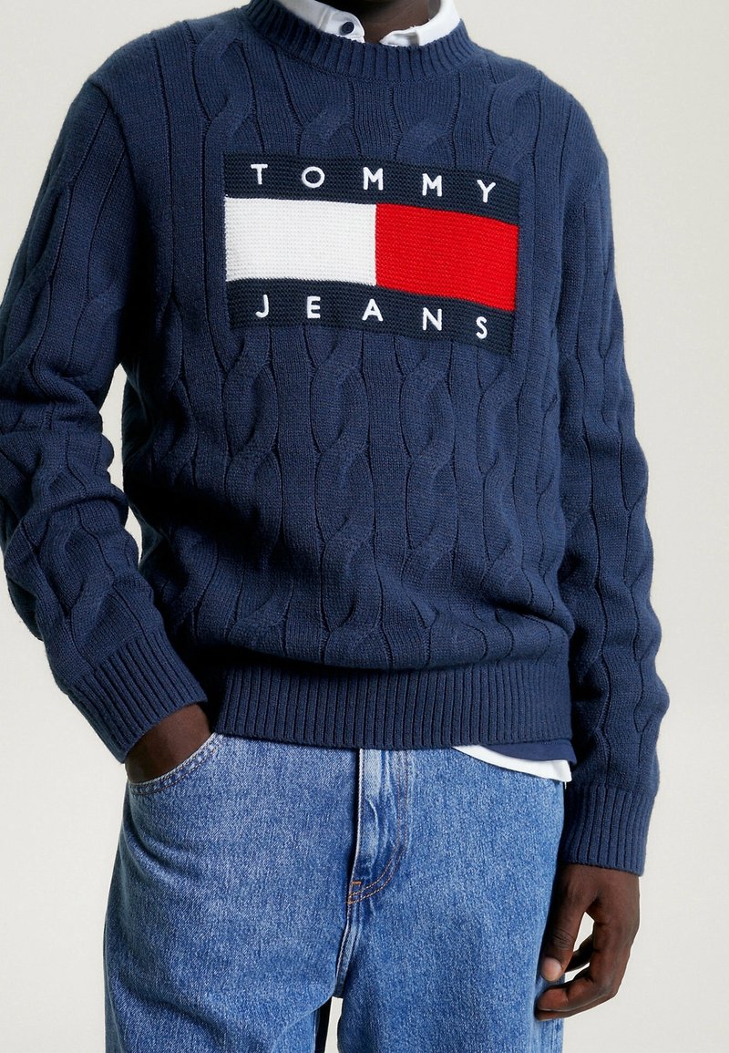 Námořnický svetr s pleteným vzorem, který má kontrastní obdélníkovou nášivku s nápisem "TOMMY JEANS" bílými písmeny a blokem červené a bílé barvy.