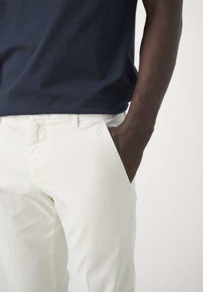 Pantaloni di cotone bianchi con una texture liscia e tasche laterali, abbinati a una maglietta blu navy. La vestibilità è rilassata e casual.