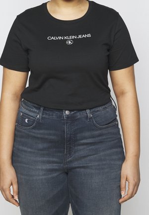 Černé triko s krátkým rukávem s bílým logem "CALVIN KLEIN JEANS", spárované s tmavě modrými džínami, které mají zip a přední kapsy.