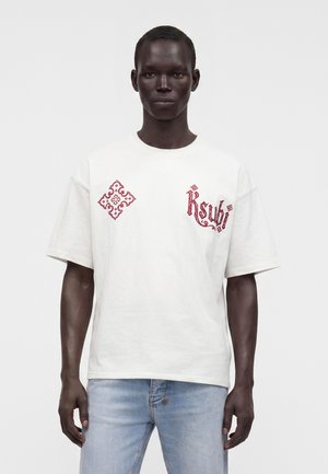 Mandlig model iført en hvid T-shirt med rød dekorativ tekst og design på brystet, kombineret med lyseblå jeans mod en ensfarvet baggrund.