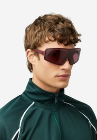 Temno bordo sončna očala z geometričnim dizajnom, temno zatemnjenimi lečami in debelim okvirjem. Nošena z temno zeleno jakno z zadrgo.