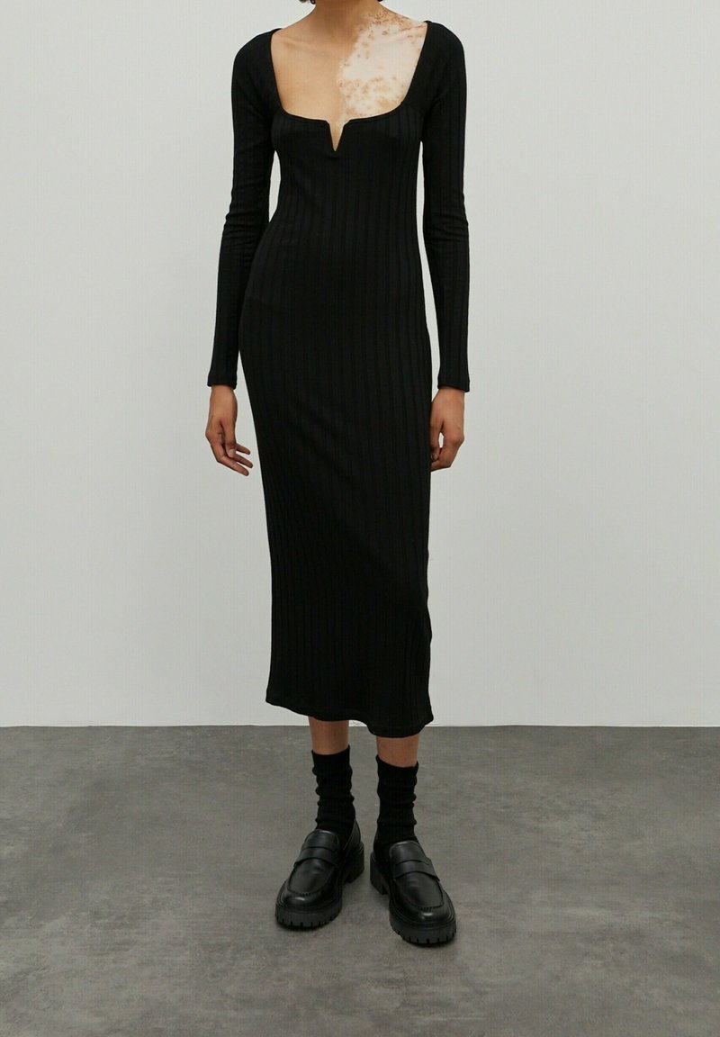 Schwarzes, geripptes Midikleid mit langen Ärmeln, Rundhalsausschnitt und subtiler V-Ausschnitt. Kombiniert mit klobigen, schwarzen Loafers und schwarzen Socken.