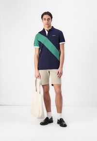 Marineblå polo med en grønn diagonal stripe, hvit krage, kombinert med beige shorts. Holder en kanvas tote bag, iført svarte loafere og hvite sokker.