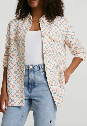 Mujer con una camisa abotonada de estampado con formas geométricas rojas y azules, botones de perla, una camiseta blanca sin mangas y jeans de tiro alto azul claro.