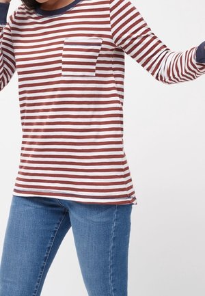 Osoba ubrana w brązowo-biały pasiastej koszuli z długim rękawem i kieszenią na piersi oraz niebieskich dżinsach na jednolitym tle.