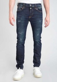 Jeans ajustados de color azul oscuro con una textura desgastada, que presentan parches rasgados y un cierre de botón. Combinados con zapatillas blancas.