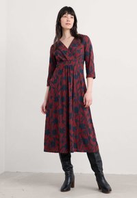 Femeie în picioare cu mâinile relaxate, purtând o rochie midi florală roșie și bleumarin, cu mâneci până la trei sferturi și botine negre cu toc.