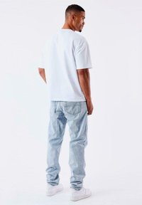 Camiseta blanca de manga corta con cuello redondo, combinada con jeans de mezclilla azul claro. Modelo vistiendo zapatillas blancas, visto desde la espalda.