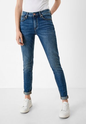 Kvinde iført opsmøgede blå skinny jeans og hvide sneakers, står med den ene arm afslappet langs siden.