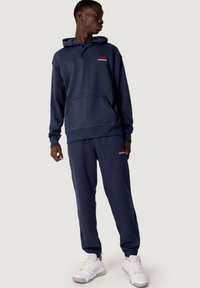 Sudadera con capucha azul marino con un bolsillo frontal, pantalones jogger a juego y logo rojo. Tejido suave, corte relajado y puños acanalados. Zapatillas blancas.