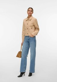 Femme portant une veste beige boutonnée, un jean bleu, des bottes à talons noires, tenant un sac à main marron sur fond blanc.
