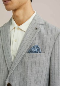 Blazer a rayas gris claro con solapa de muesca, detalle de botones y un pañuelo de bolsillo azul con estampado asomando del bolsillo del pecho. Polo blanco debajo.