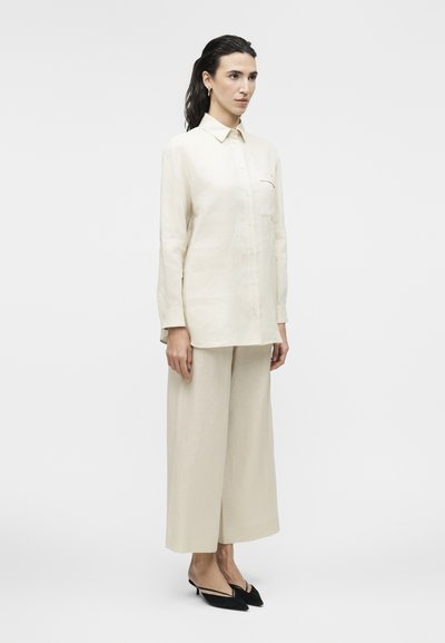 Γυναίκα στέκεται φορώντας κρεμ πουκάμισο με μακριά μανίκια και κουμπιά, παντελόνι με φαρδιά μπατζάκια στο ίδιο χρώμα και μαύρες μυτερές γόβες με λουράκι στο πίσω μέρος, σε λευκό φόντο.