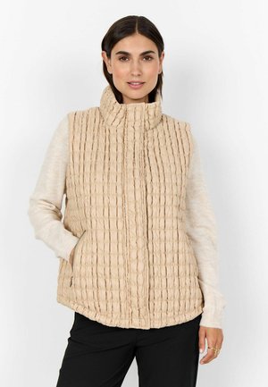 Beige kviltad väst med hög krage, med vertikala kanaler, dragkedjefickor och en texturerad yta. Bärs över en ljus tröja.
